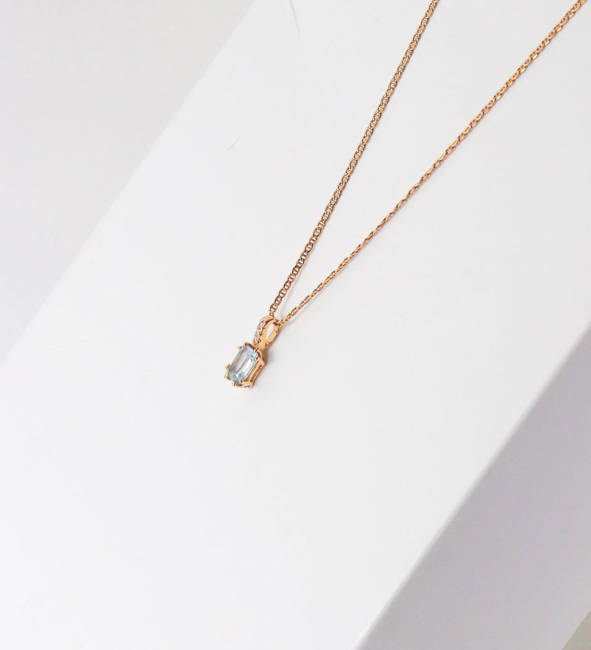 Gold pendant necklace with a pale blue stone arranged on a white Betonvton display block.