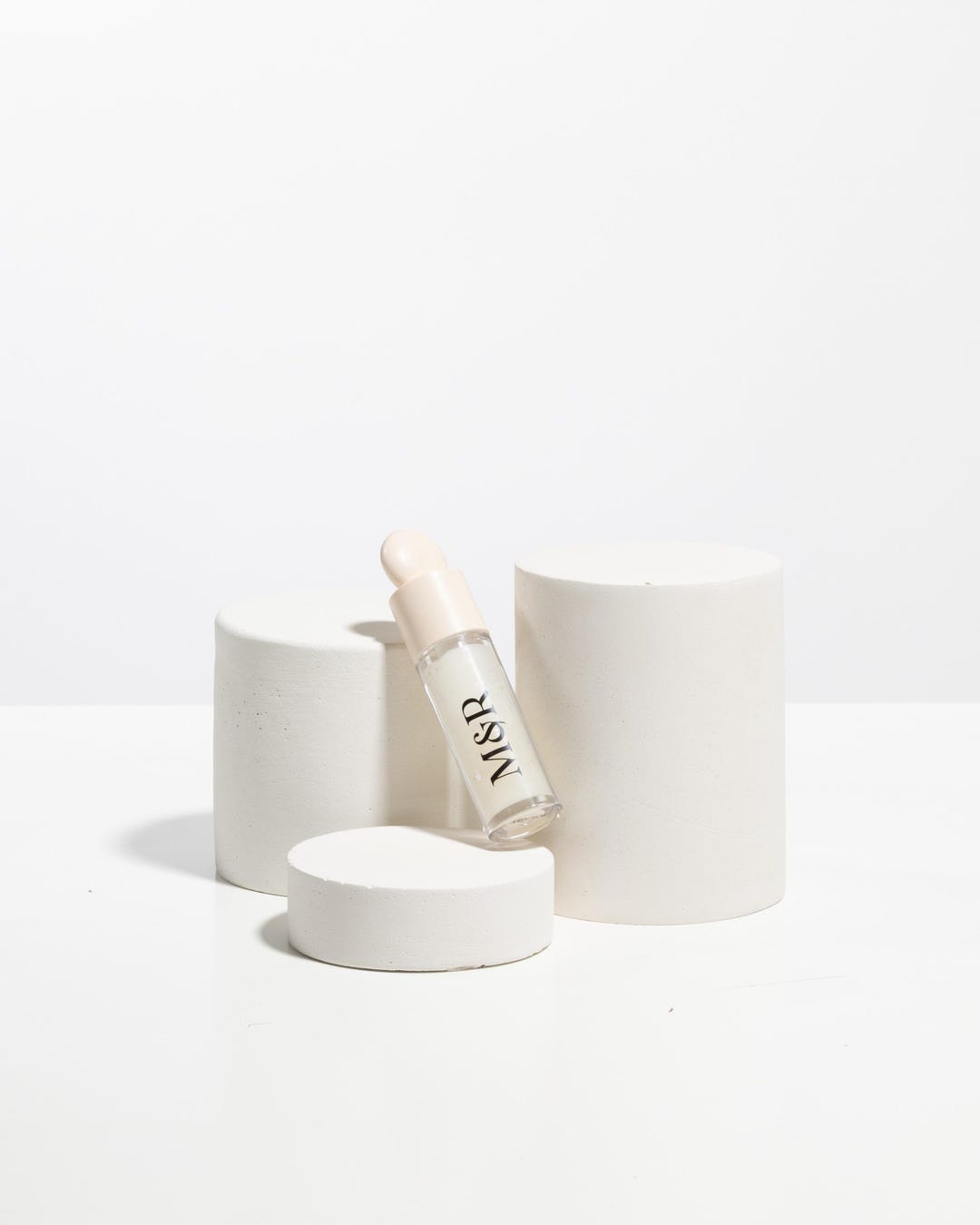 Cosmetic dropper bottle styled between matte white cylinder display pedestals